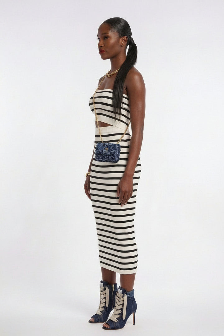 A model wearing a black and white horizontally striped sleeveless midi dress with a cutout at the waist, styled with a blue patterned crossbody bag and blue lace-up ankle boots against a plain white studio background.