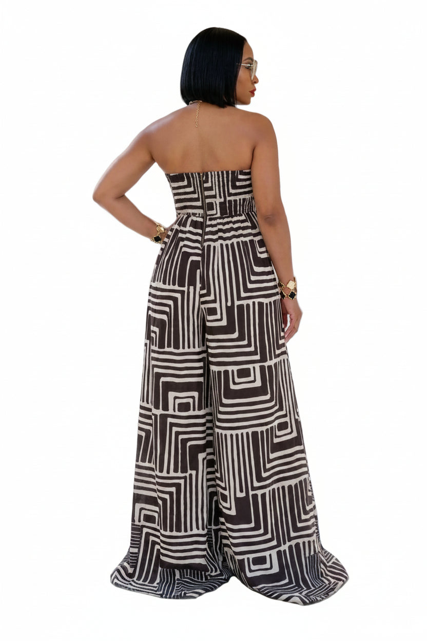 A woman stands in a grand classical hall wearing a strapless wide-leg jumpsuit with a bold black and off-white geometric maze-like pattern, shown from the back.