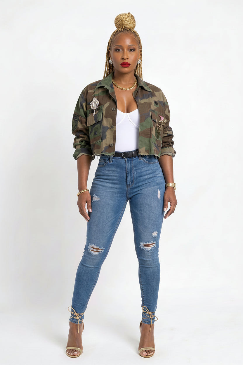 A woman modeling a cropped camouflage jacket over a white tank top, paired with high-waisted blue distressed skinny jeans and lace-up ankle boots, against a plain white studio background.