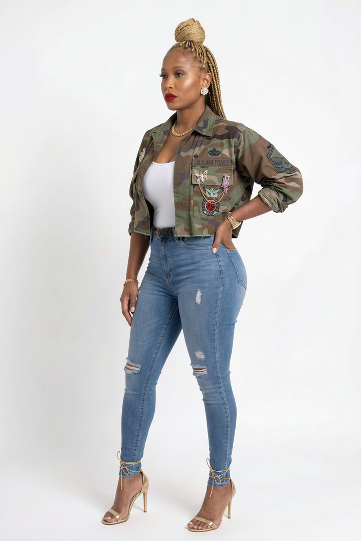 A woman modeling a cropped camouflage button-up jacket with colorful embroidered patches, worn over a white tank top and paired with distressed blue skinny jeans and nude high-heeled sandals, against a plain white studio background.